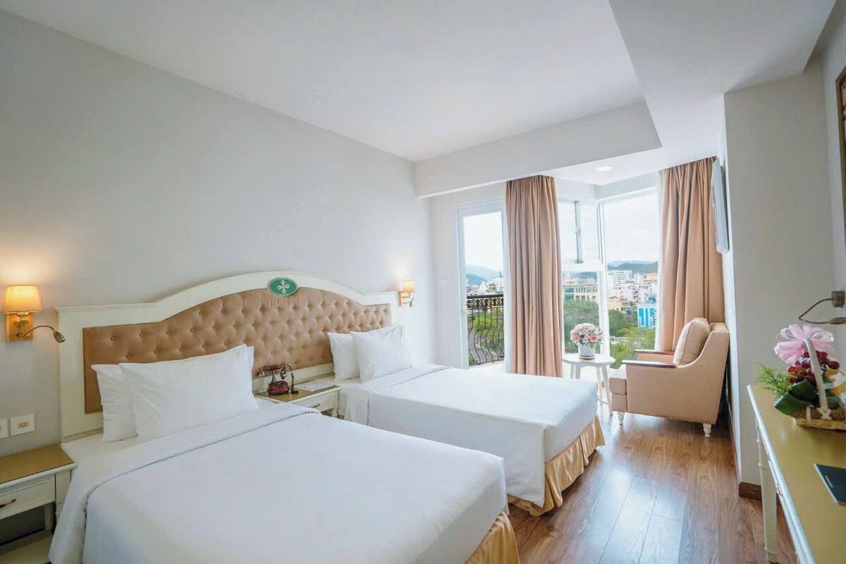 Hotelkamer met twee bedden, een groot raam met gordijnen en uitzicht op de stad, plus een klein tafeltje met bloemen.