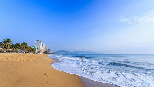A beach with a sandy shore, ocean waves, and tall buildings in the background. - Nha VIET Nha Trang Hotel (Nha Trang)