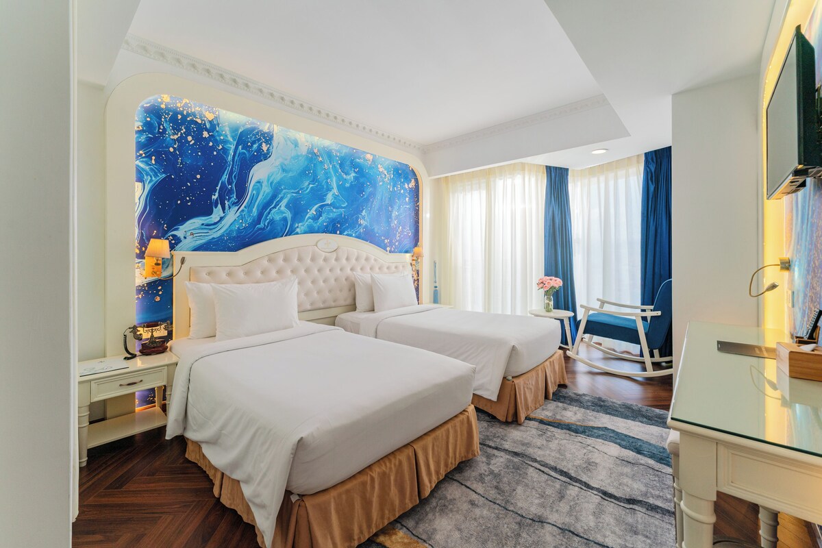 Ein Hotelzimmer mit zwei Betten, einer großen Wandmalerei, einem Schreibtisch mit Lampe und einem Fernseher an der Wand.