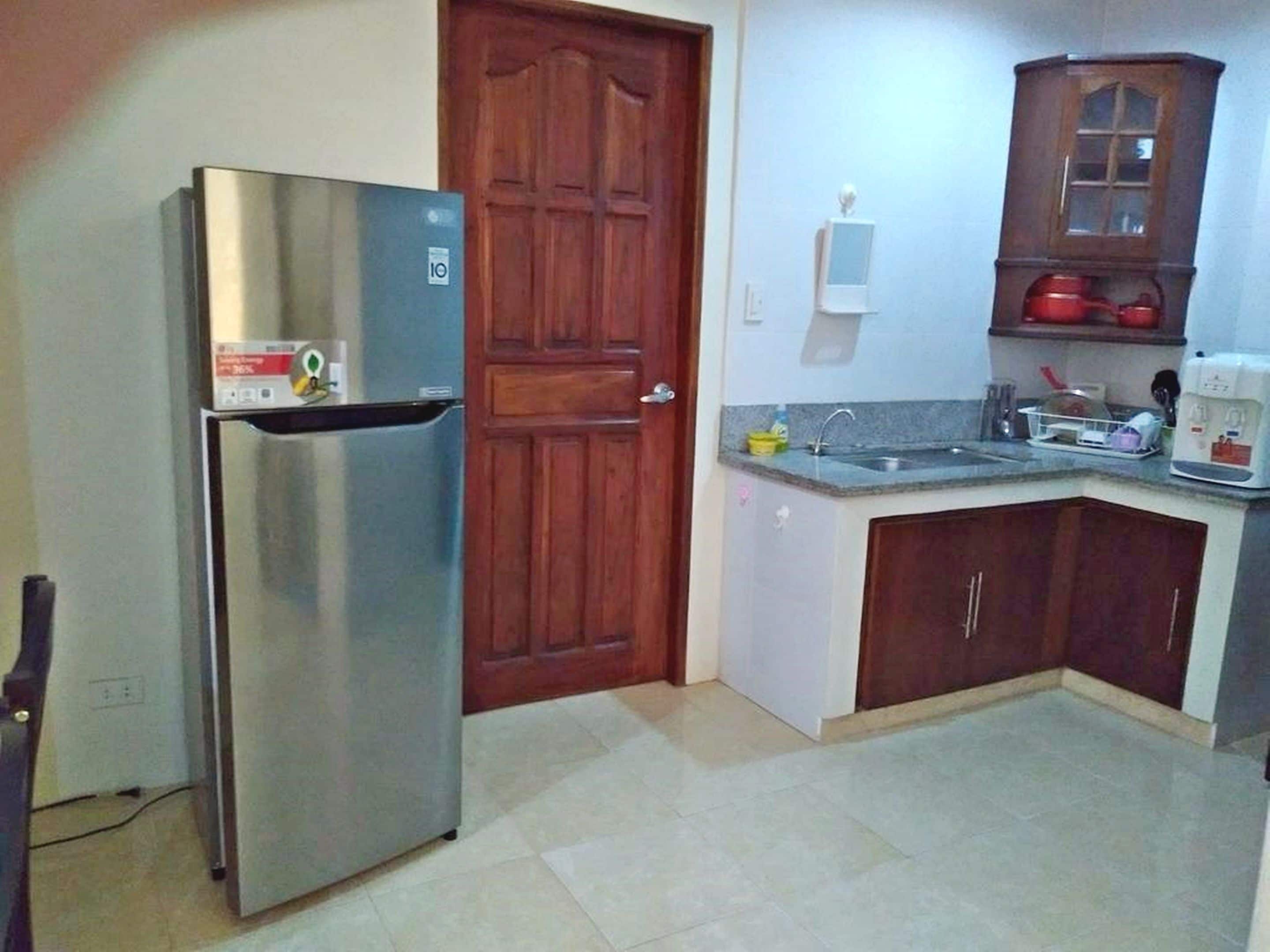 Una cocina compacta con un refrigerador de acero inoxidable, una puerta de madera, un lavamanos empotrado y una pequeña encimera con un hervidor.