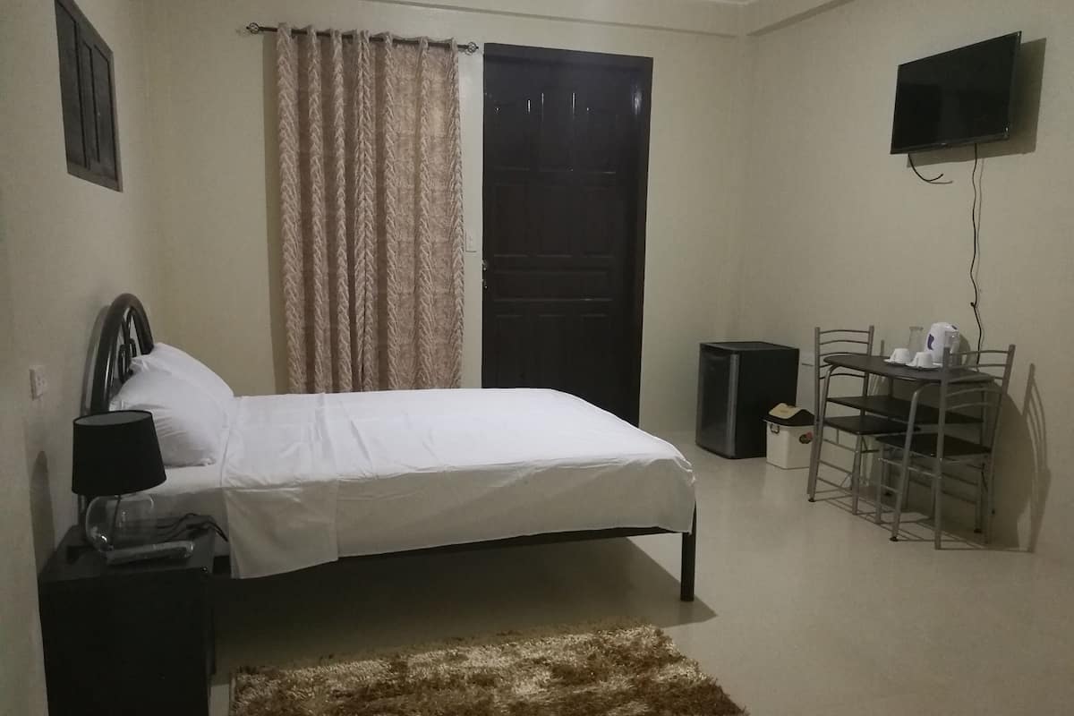 Un dormitorio con cama, escritorio, silla, televisor y una mesita con tazas y hervidor.