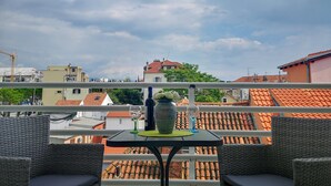 Un balcon agrémenté de meubles en osier, une table avec un vase et des verres à vin, et une vue sur les toits et les bâtiments aux tuiles rouges.