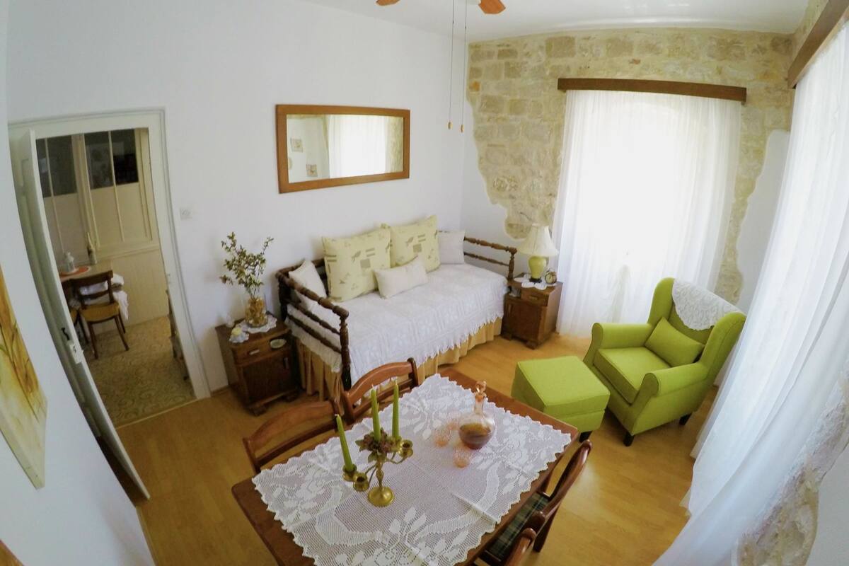Quarto com uma cama, uma poltrona verde, uma mesa de madeira com toalha rendada e um ventilador de teto.