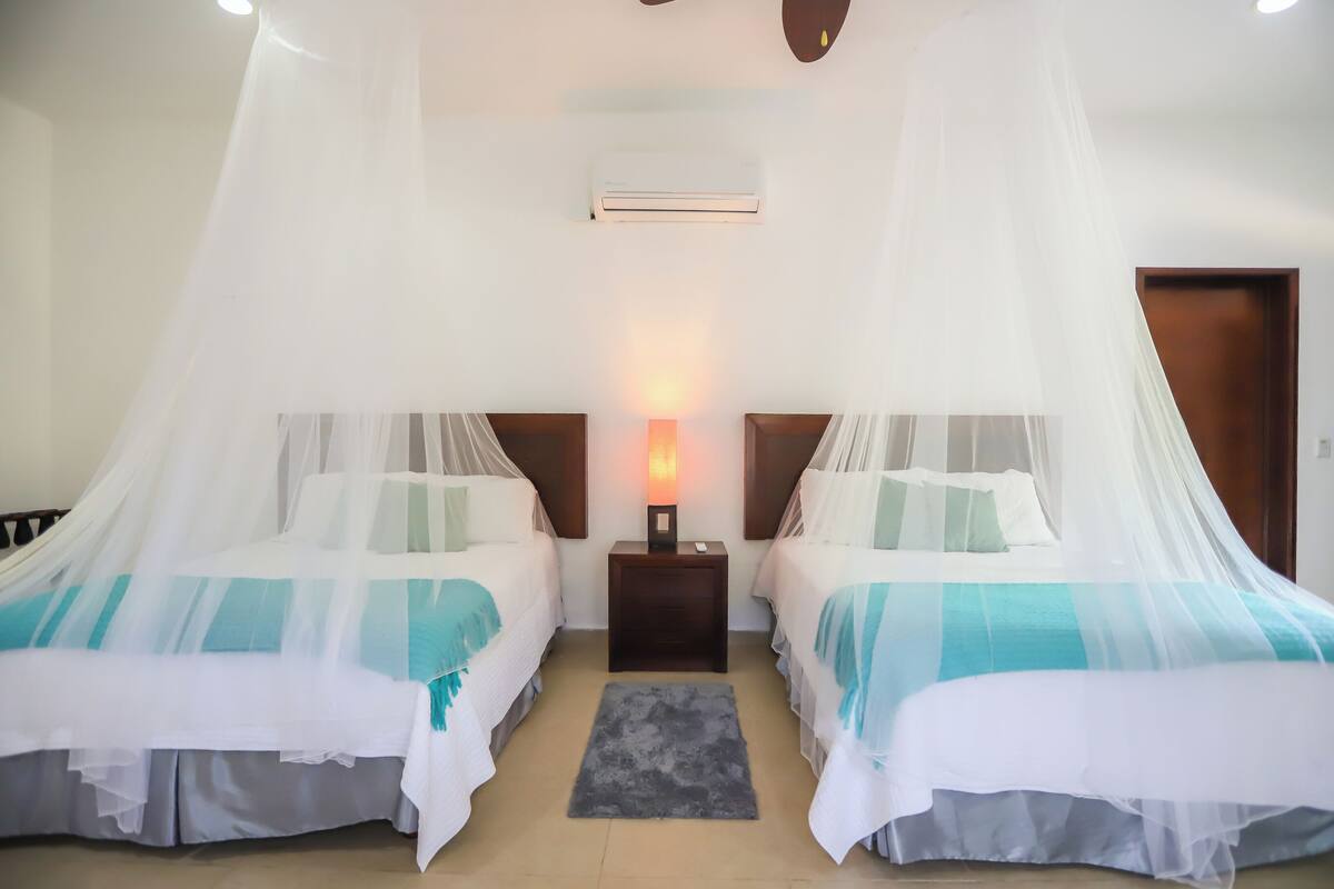 Ein Hotelzimmer mit zwei Betten, jeweils mit Baldachin, einem Nachttisch mit Lampe sowie einem kleinen Teppich auf dem Boden.