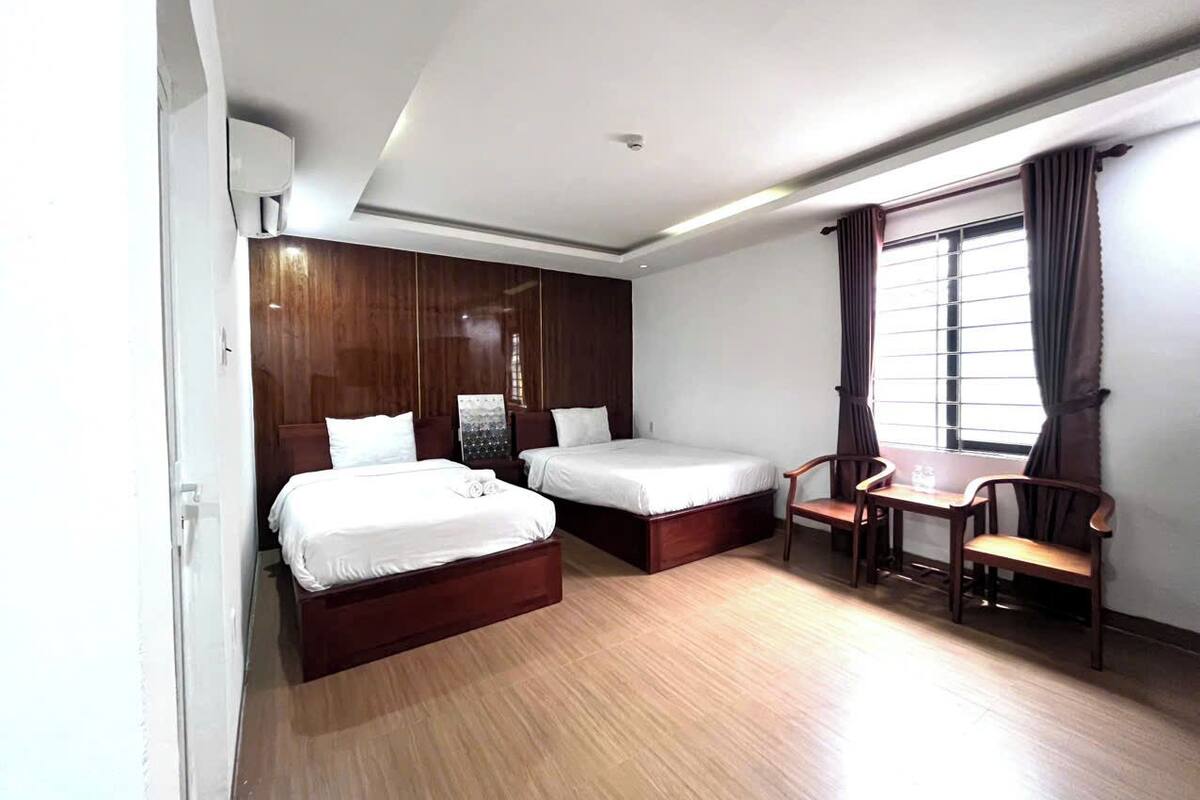 A hotel room with two beds, a wooden floor, a desk with chairs, and a window with curtains.