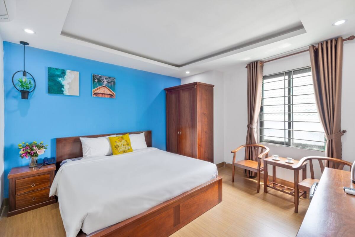A bedroom with a bed, wooden nightstands, a desk with chairs, a wardrobe, and a window with curtains.