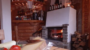Un interior acollidor amb una llar de pedra, prestatges de fusta amb ampolles, i una taula de tall amb formatge i tomàquets.