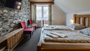 Habitació amb paret de pedra, sostre de fusta, llit amb roba de llit estampada, una cadira vermella, un armari de fusta i un televisor fixat a la paret.