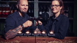 Zwei Personen sitzen an einem Tisch mit angezündeten Kerzen und Weingläsern, im Hintergrund Regale voller Weinflaschen.