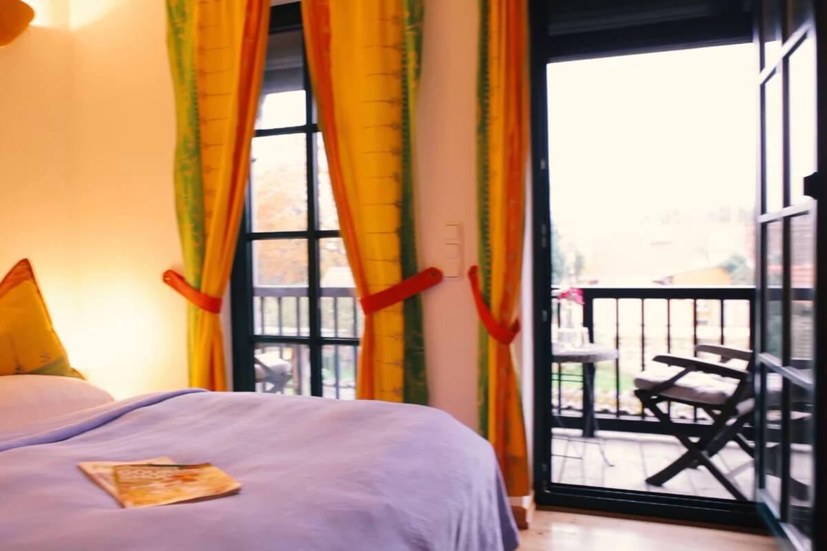A room with a bed, a window with curtains, and a balcony with a chair.