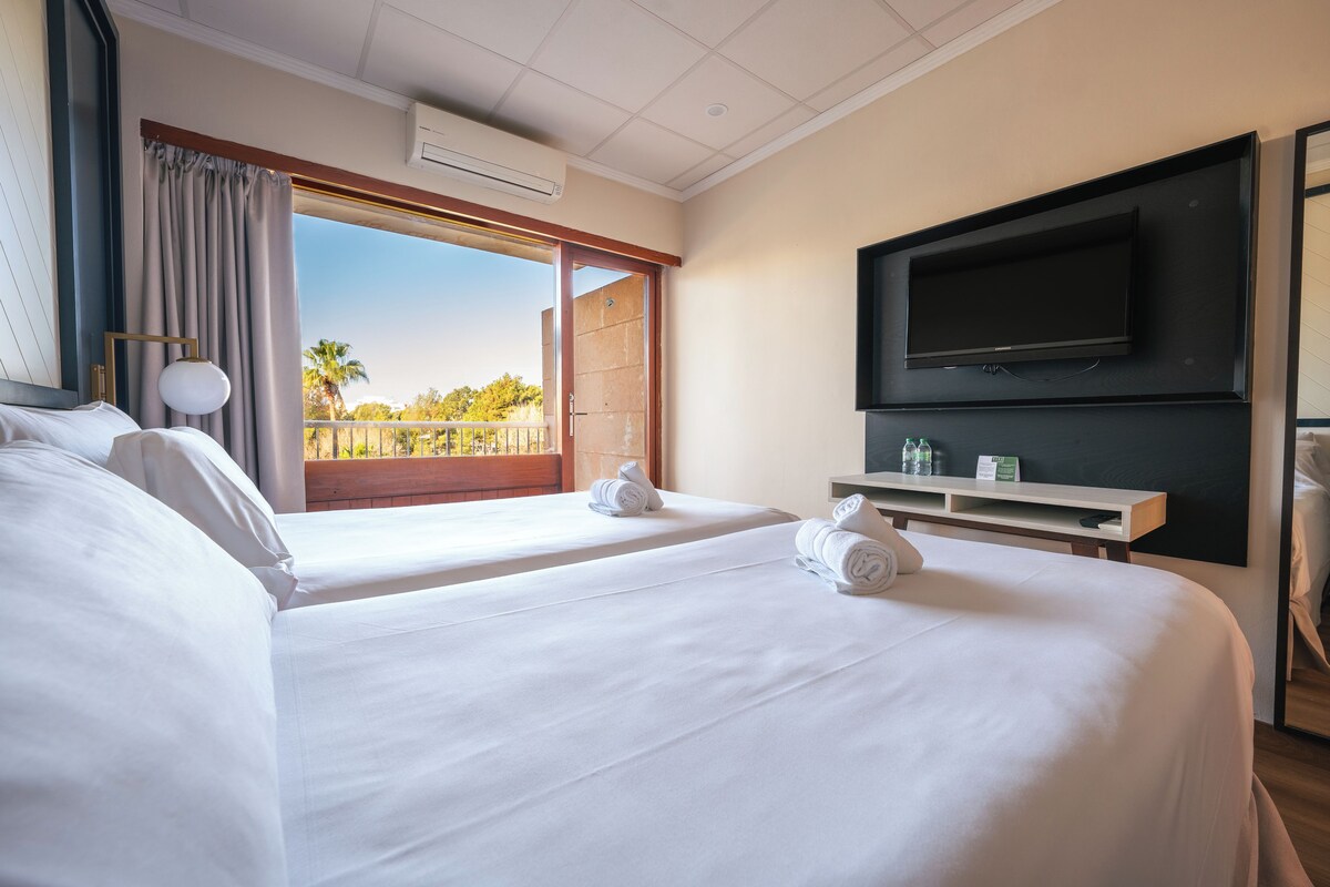 Habitación de hotel con dos camas, televisión, balcón y vista a palmeras, además de ventilador de techo.