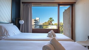 Habitación de hotel con dos camas, un balcón que ofrece vistas a edificios y palmeras, y una ventana con cortinas.