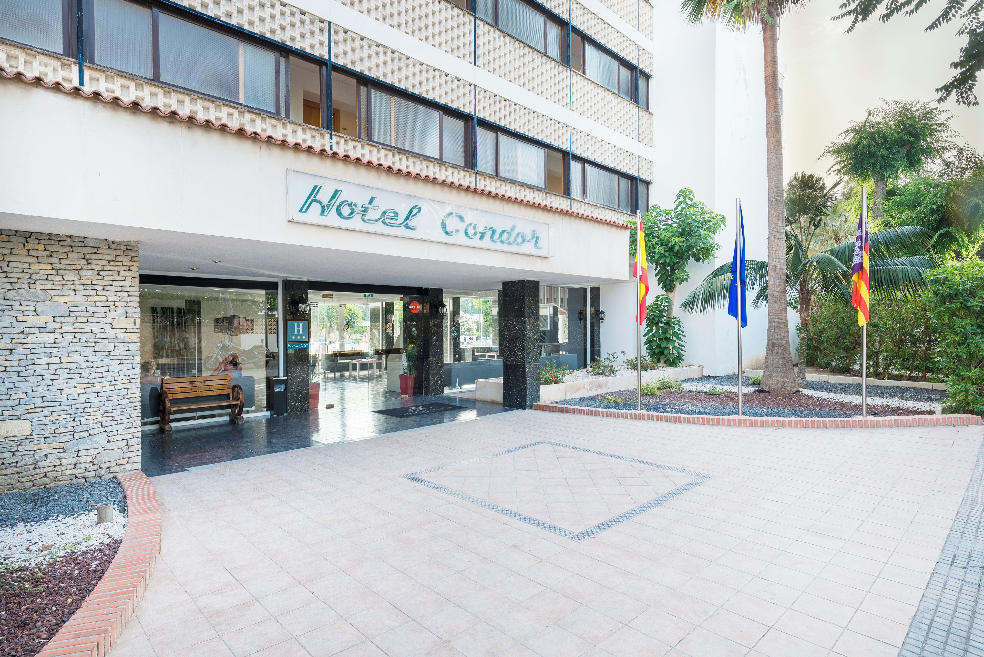 Entrada de un hotel con un camino de baldosas, un banco y un cartel que dice 'Hotel Condor'.