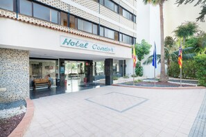 Entrada de un hotel con un camino de baldosas, un banco y un cartel que dice 'Hotel Condor'.