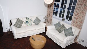Una sala de estar con dos sofás blancos, un otoman redondo de madera, una lámpara y una ventana con cortinas.