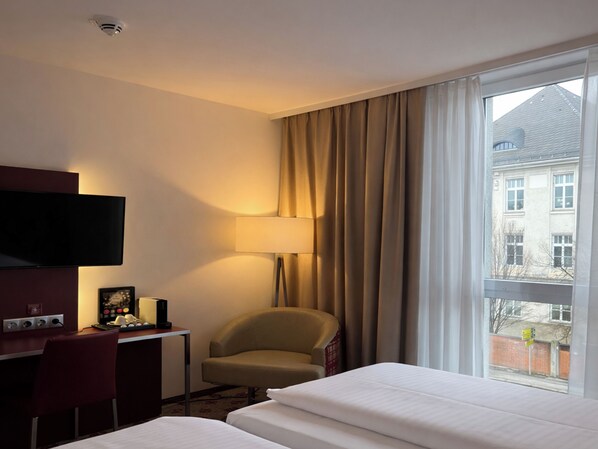 A hotel room with a bed, a desk with a TV, a chair, a lamp, and a window with a view of buildings.
