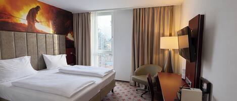 A hotel room with a large bed, a desk with a chair, a television, and a window with curtains.