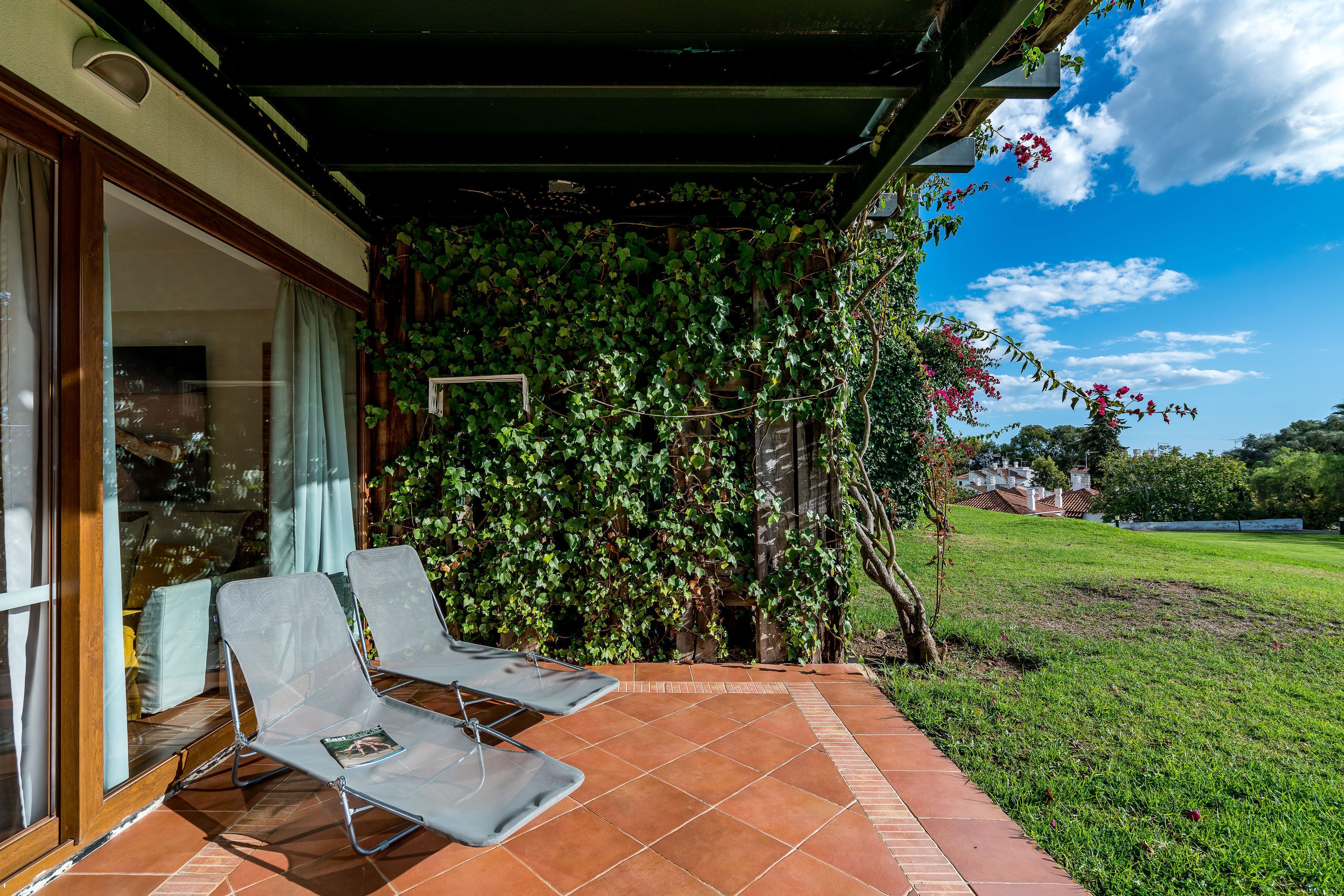 Un patio cubierto con dos sillones, suelo de baldosas y una pared verde exuberante.
