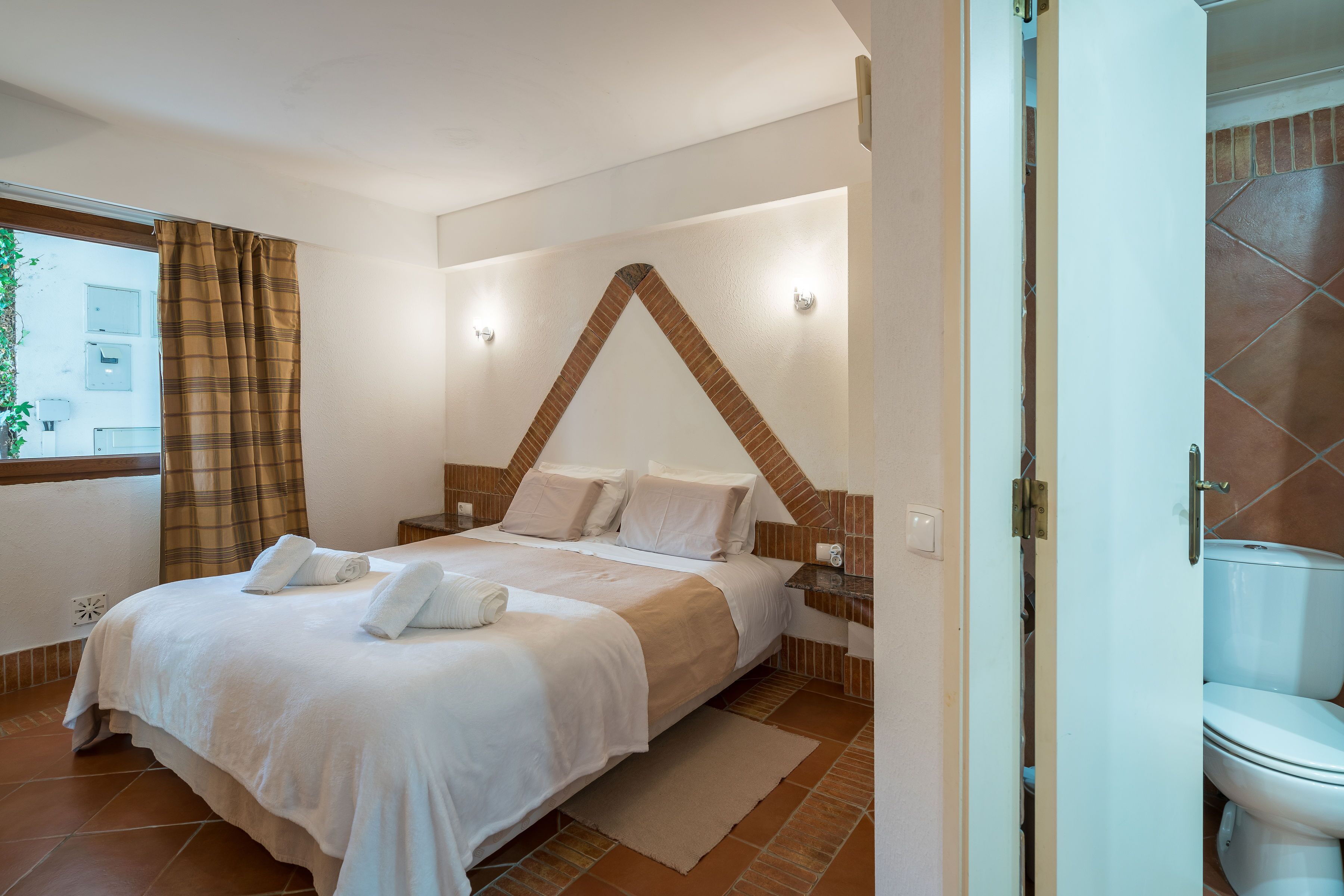 A bedroom with a bed, a nightstand, a window with curtains, and a view of the outside.