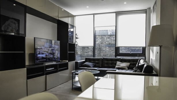 A modern living room with a large flat-screen TV, a leather sofa, a coffee table, and a dining area with a white table and chairs. - MD Luxury Apartment (London)