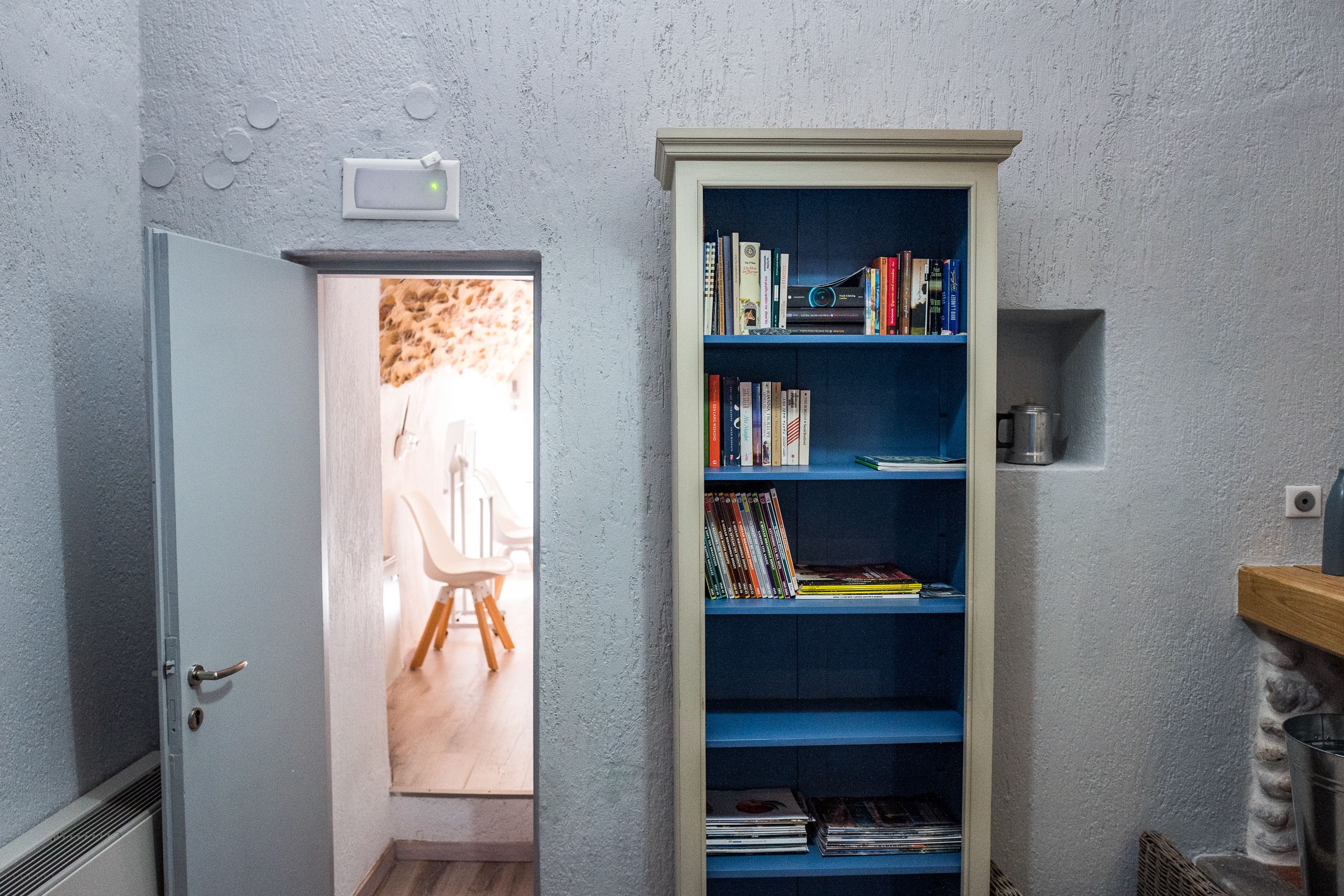 Un couloir étroit avec une bibliothèque, une porte donnant sur une pièce où se trouve une chaise, et une petite étagère avec un récipient.