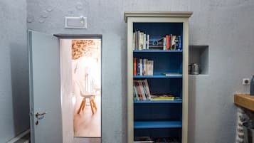Un couloir étroit avec une bibliothèque, une porte donnant sur une pièce où se trouve une chaise, et une petite étagère avec un récipient.