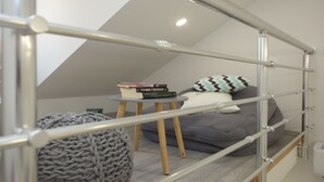 A loft-style bedroom with a sloped ceiling, a metal railing, a gray sofa, a small round ottoman, and a stack of books. - Central Old Town Rooms (Split)