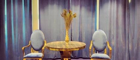 Una habitación con una mesa redonda y dos sillas, un florero con flores secas y cortinas oscuras.