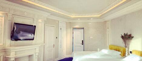 A hotel room with a large bed, a flat-screen TV mounted on the wall, a fireplace, and a door leading to another room.