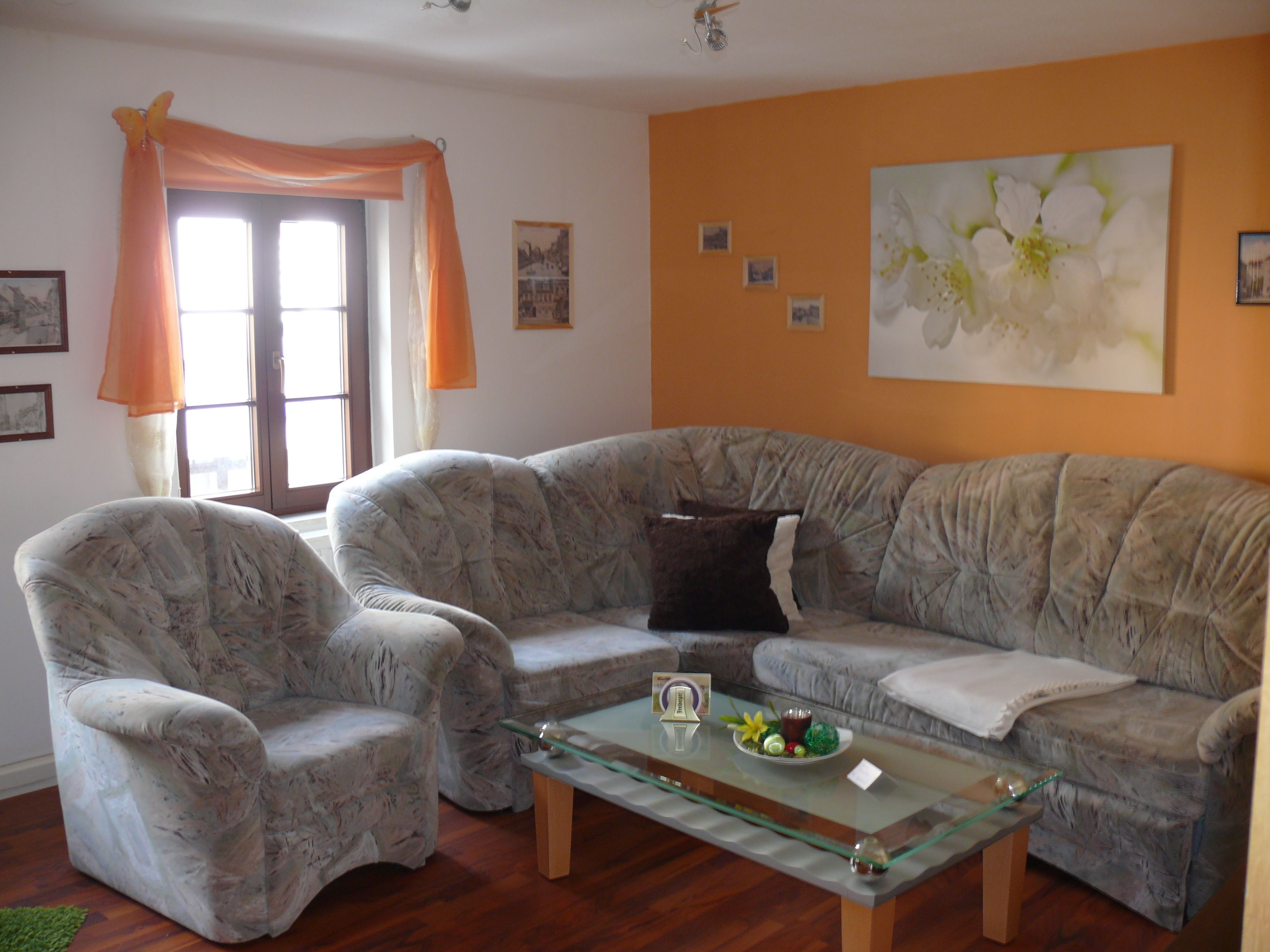 A living room with a grey sofa, two armchairs, a glass coffee table, and a window with orange curtains.