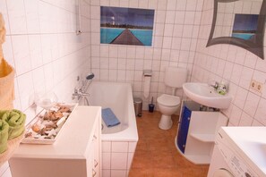 Un baño con bañera, inodoro, lavamanos y una pequeña ventana con vista al mar.