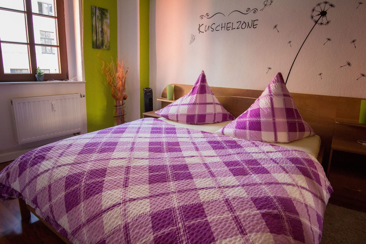 Quarto com colcha quadricula roxa e branca, parede verde e janela com vista.