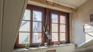 Habitación con ventana que da a un edificio de techo con tejas. Hay plantas en macetas en el alféizar y un cuadro en la pared.