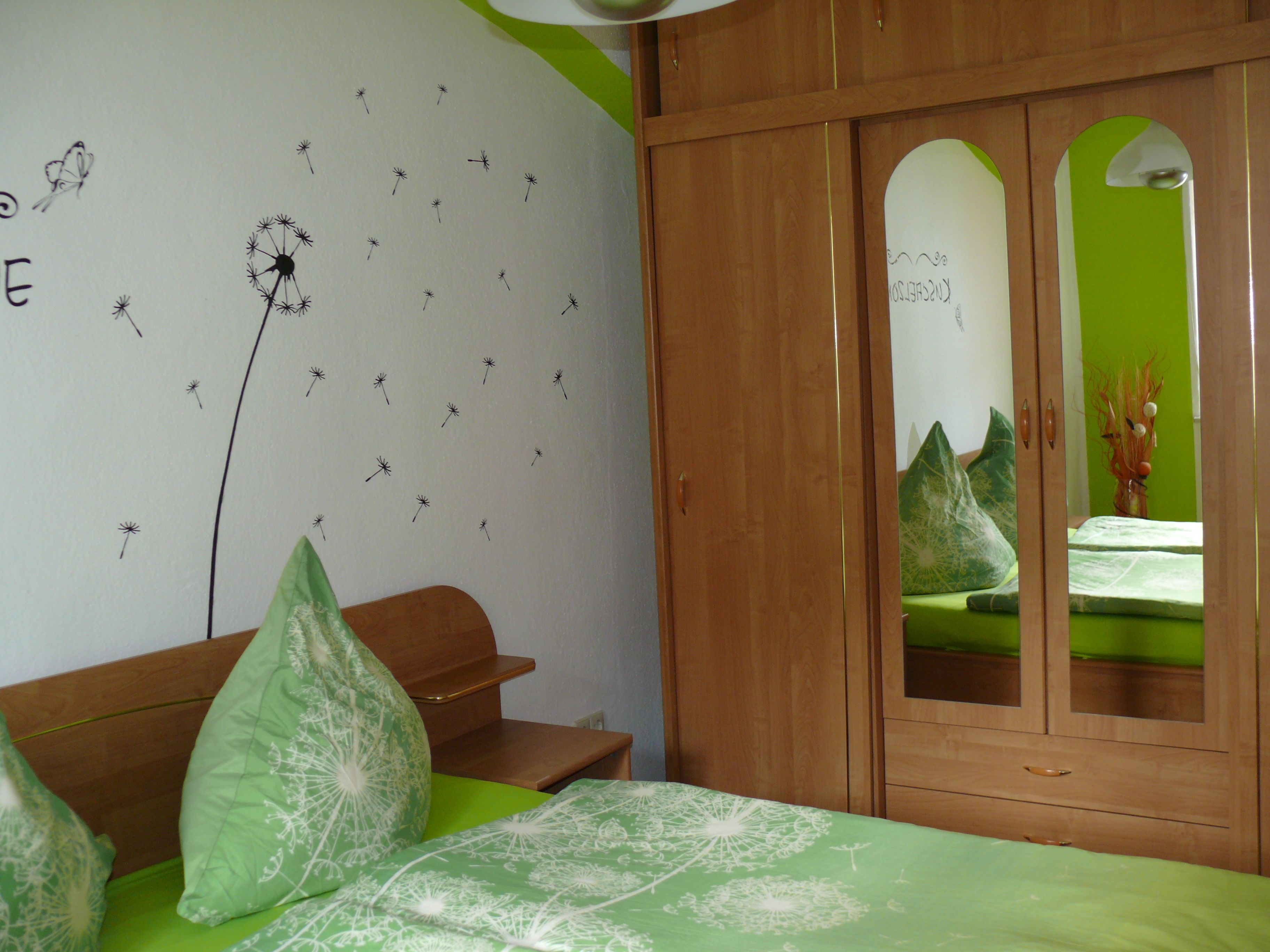 A bedroom with a green bedspread, a wooden wardrobe, and a wall with a dandelion pattern.