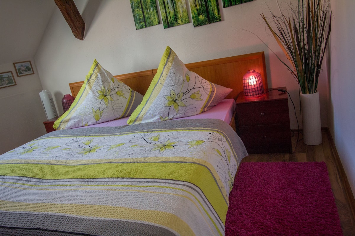 Cama bem arrumada com roupa de cama floral, luminária de cabeceira e um vaso com plantas altas.