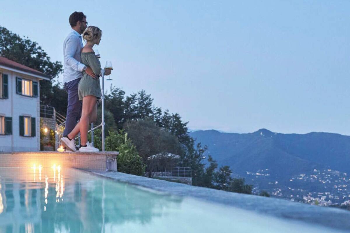 Una pareja está junto a una piscina con vistas a una montaña y una casa al fondo.