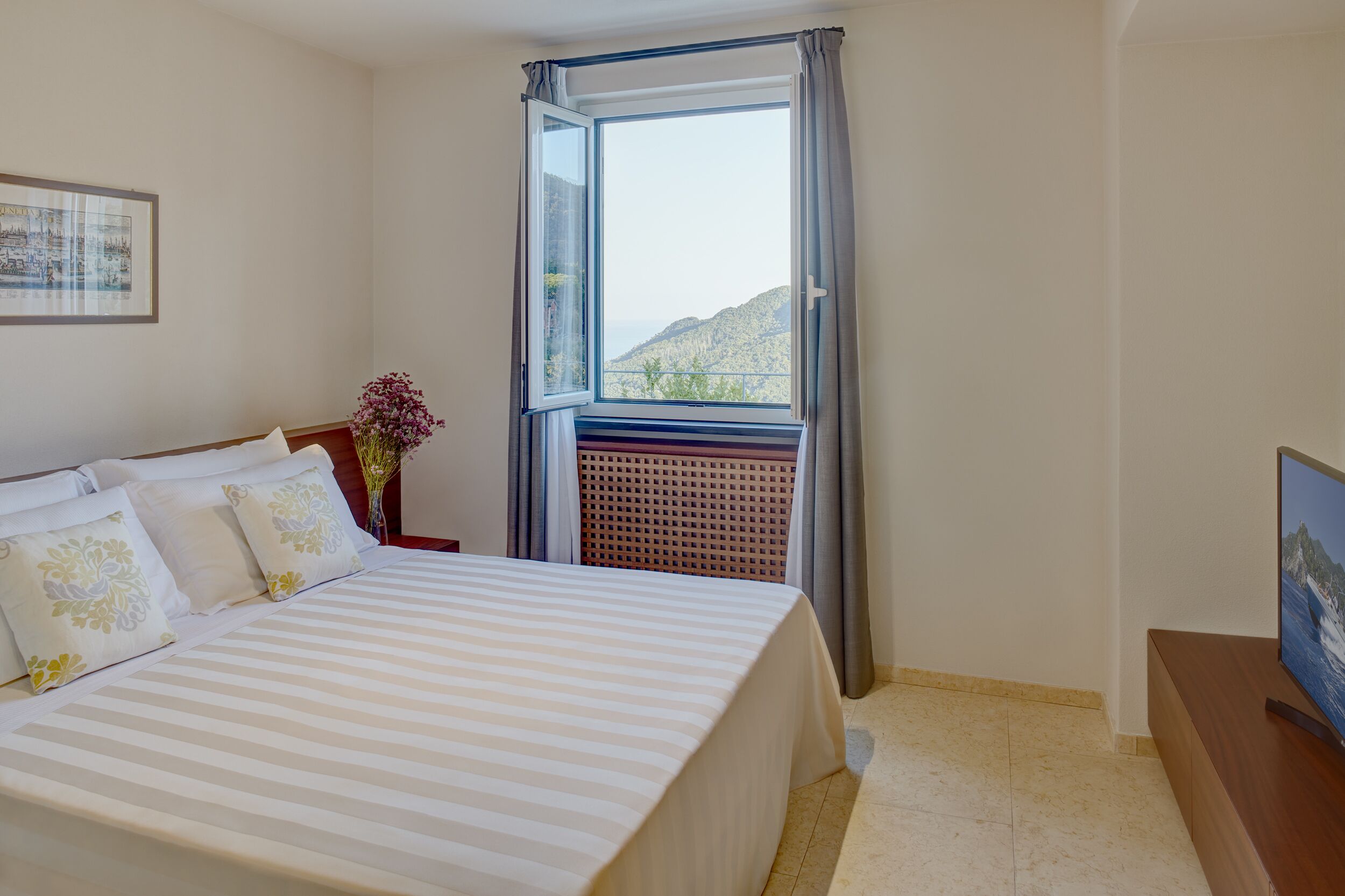 A hotel room with a bed, a TV, a window with a view of mountains, and a framed picture on the wall.