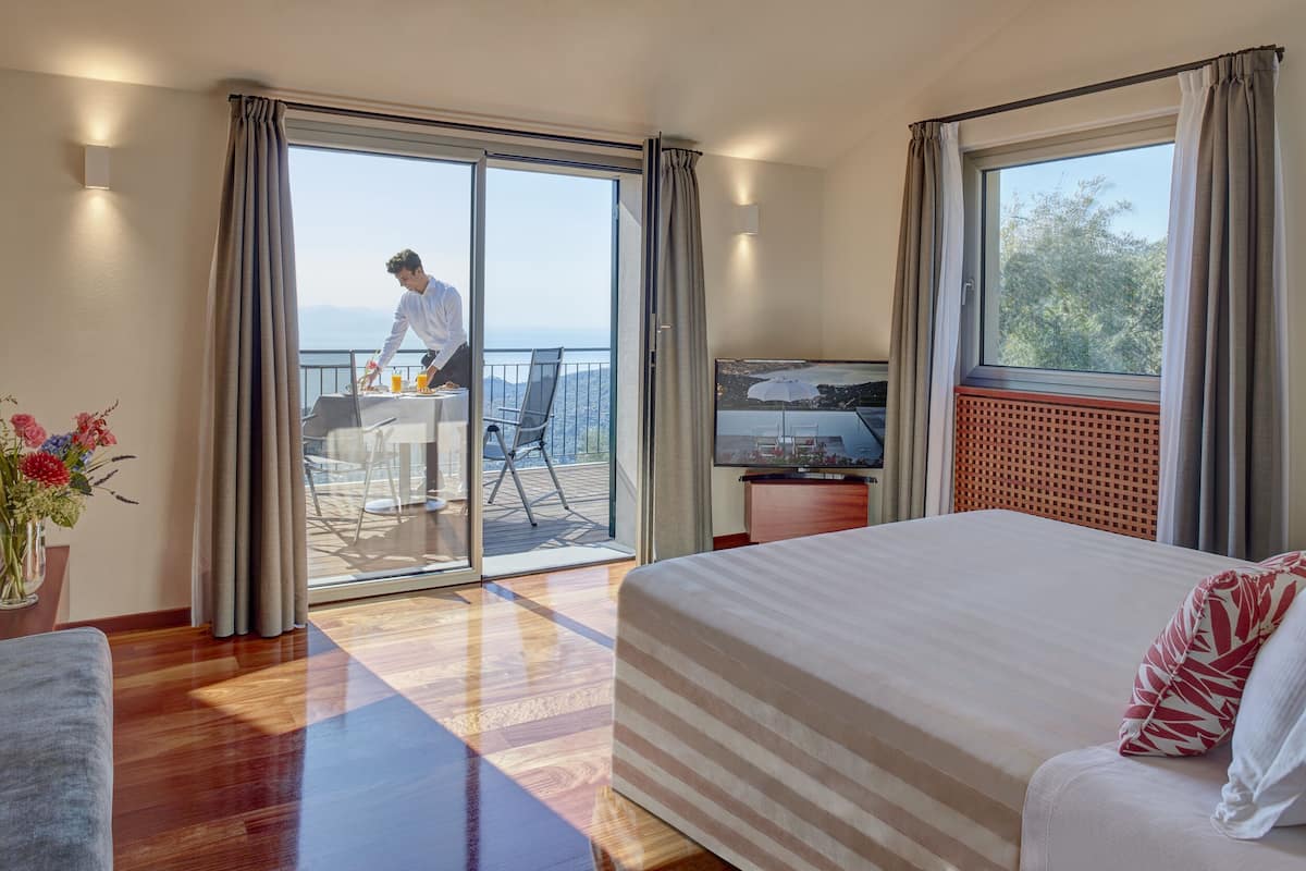 Habitación de hotel con una cama grande, televisión, balcón con mesa y sillas, y vista al mar.