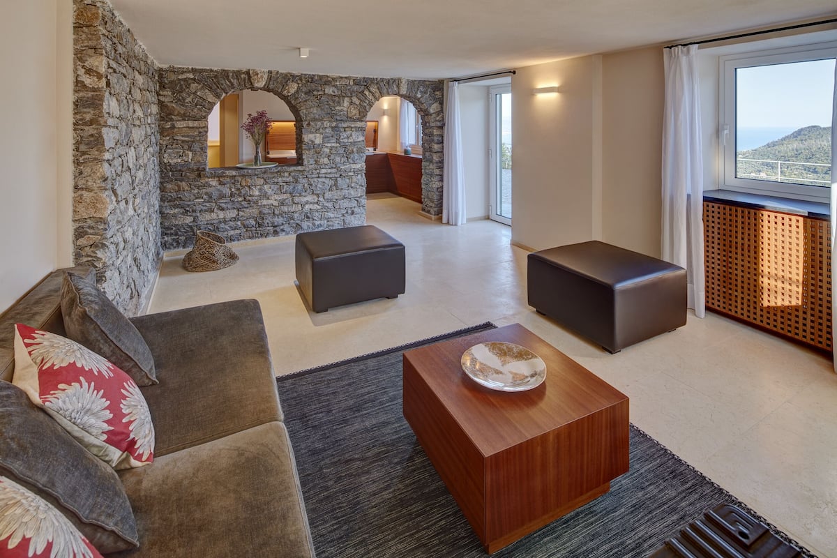 Una sala de estar moderna con paredes de piedra, un sofá, dos otomanas, una mesa de centro de madera y vistas a un balcón.