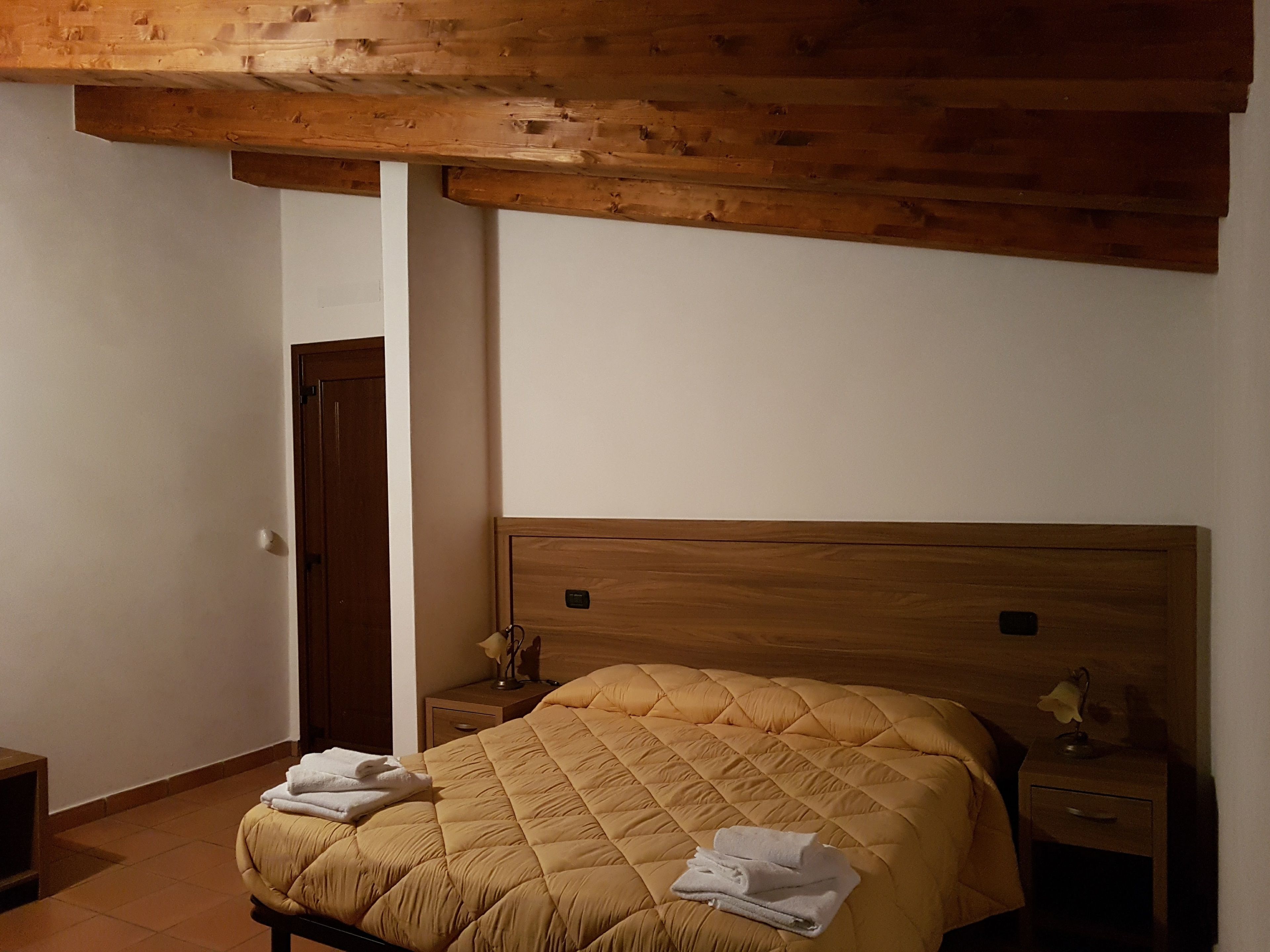 Une chambre à coucher avec une tête de lit en bois, un lit recouvert d’une courtepointe, une table de chevet avec une lampe, et une porte visible au fond.