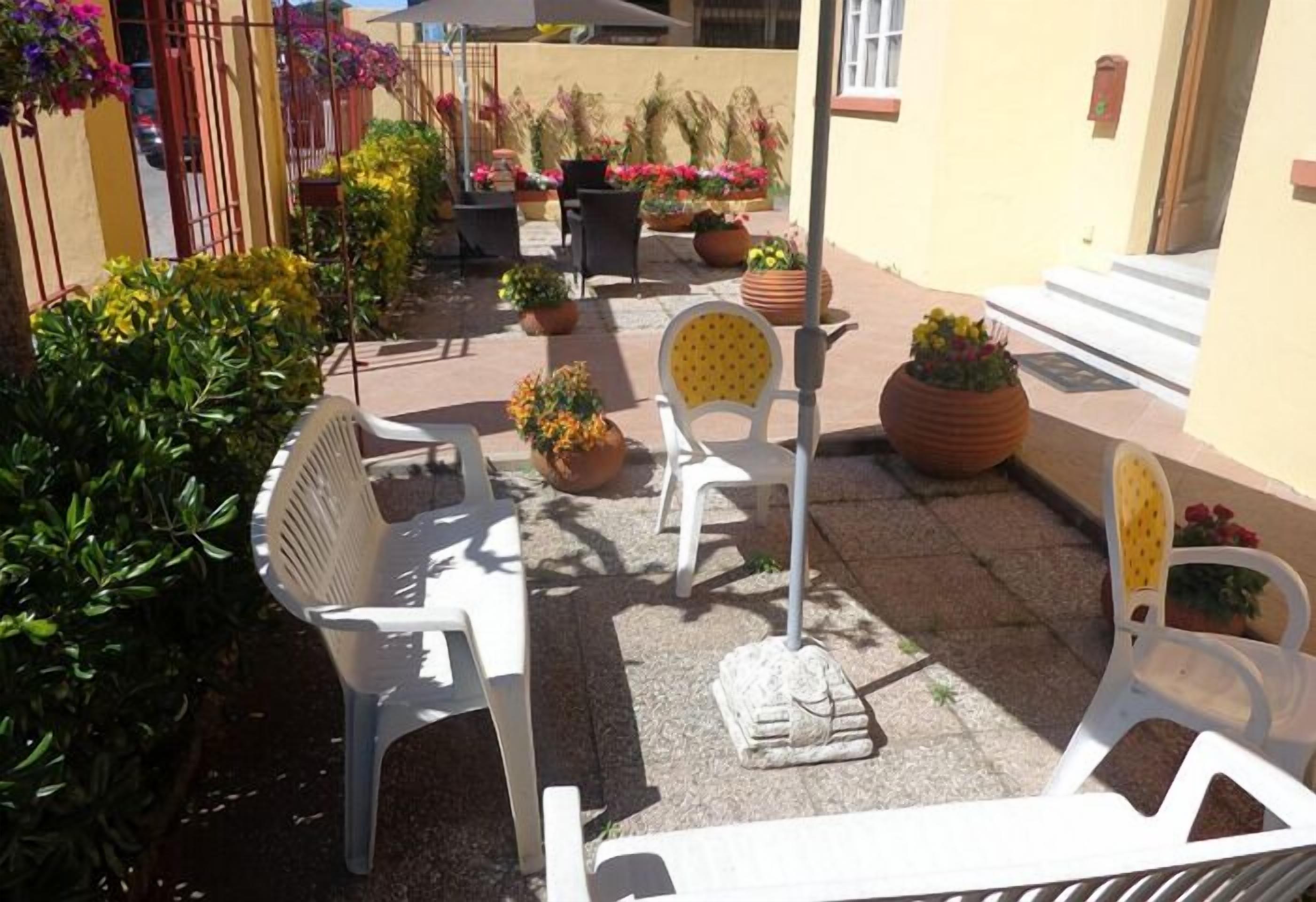 Un'area patio all'aperto con sedie di plastica bianca, piante in vaso e una facciata di un edificio gialla.