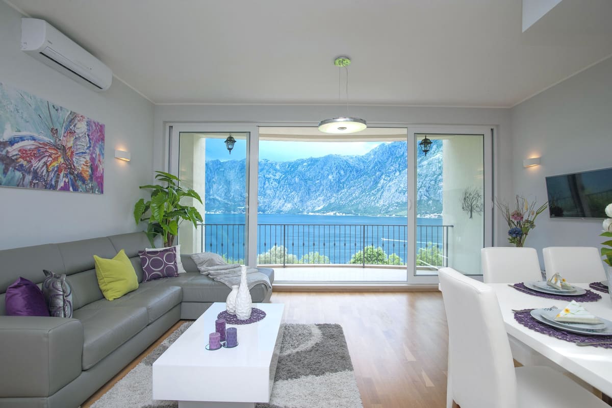 Uma sala de estar moderna com um sofá cinzento, uma mesa de centro branca e uma área de jantar com uma mesa posta para quatro pessoas. Uma grande janela oferece vista para montanhas cobertas de neve e um lago.