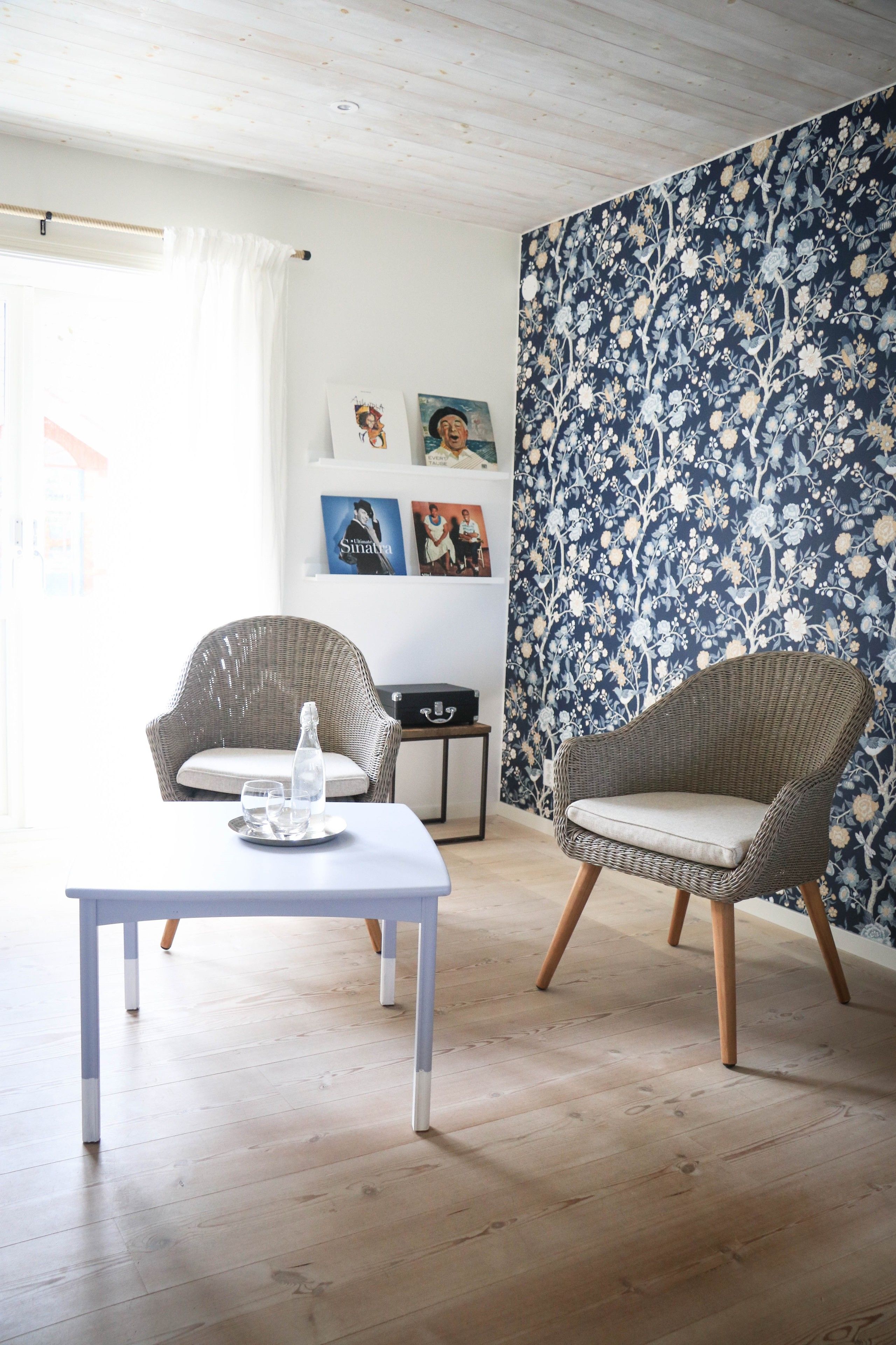 Una habitación con dos sillones de mimbre, una mesa blanca con una jarrón de vidrio y vasos, y un papel tapiz floral.