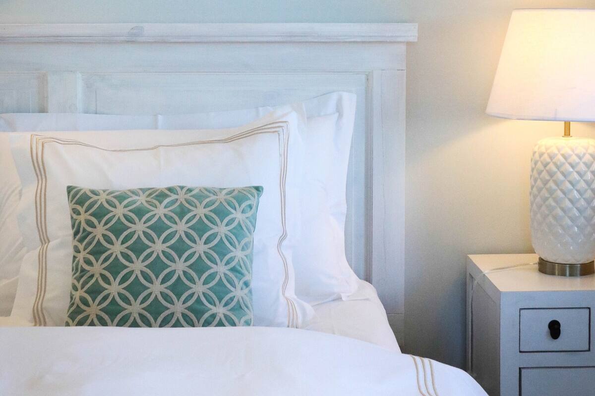 A neatly made bed with white bedding and a patterned pillow, a bedside lamp, and a wall-mounted branch decoration.
