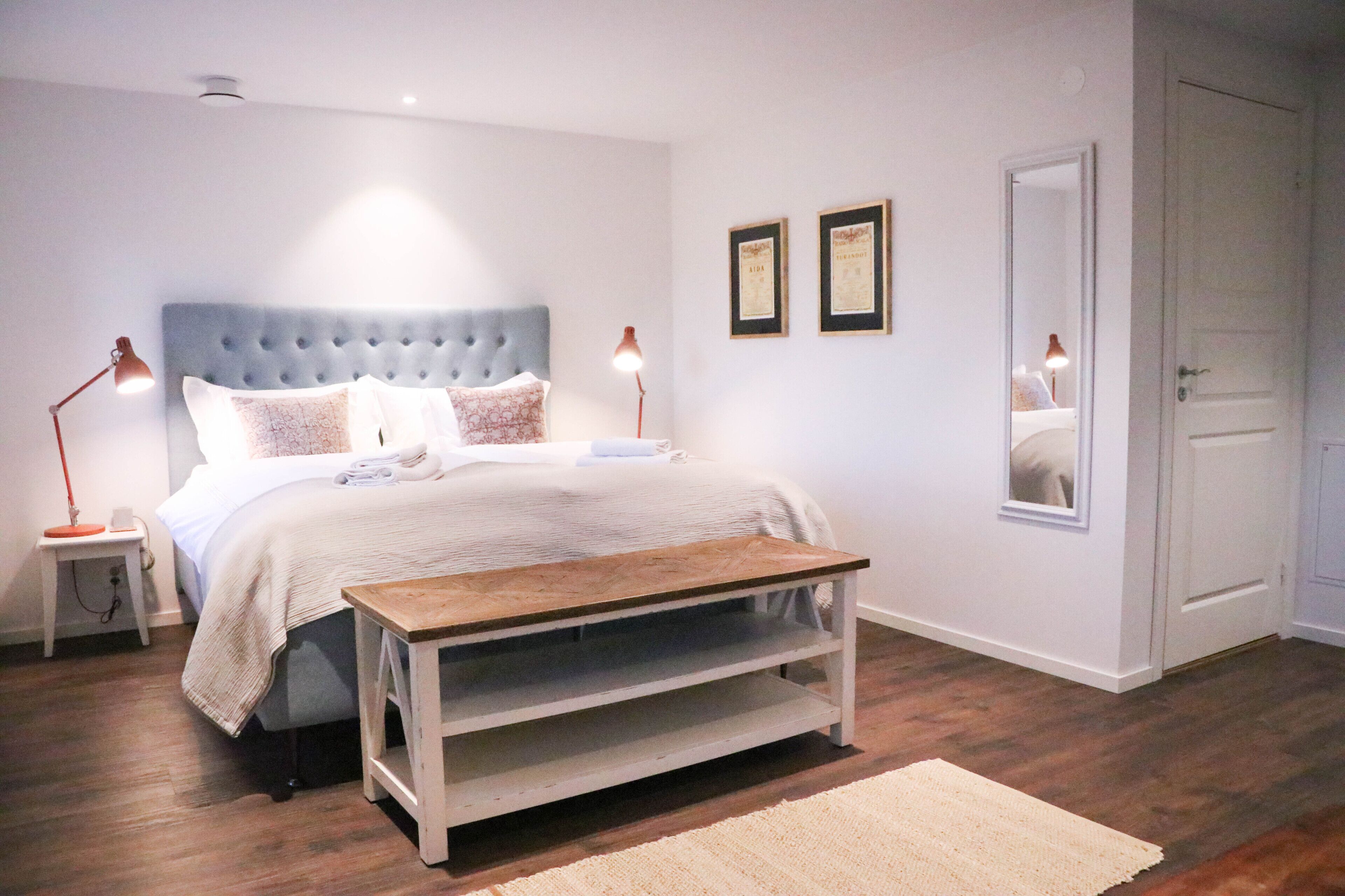 A bedroom with a bed, bedside lamps, a nightstand, a wooden bench, a rug, and a door.