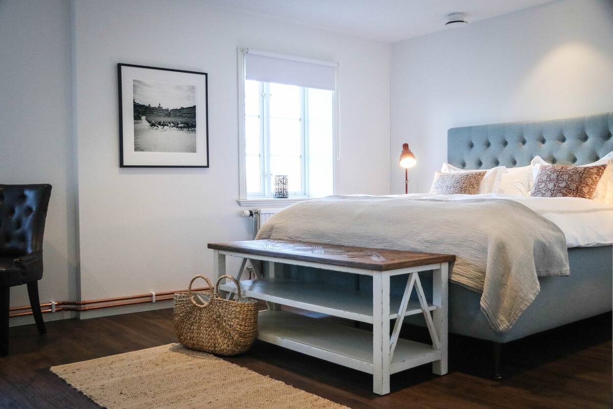 A bedroom with a bed, a chair, a bedside table, a lamp, and a framed picture on the wall.