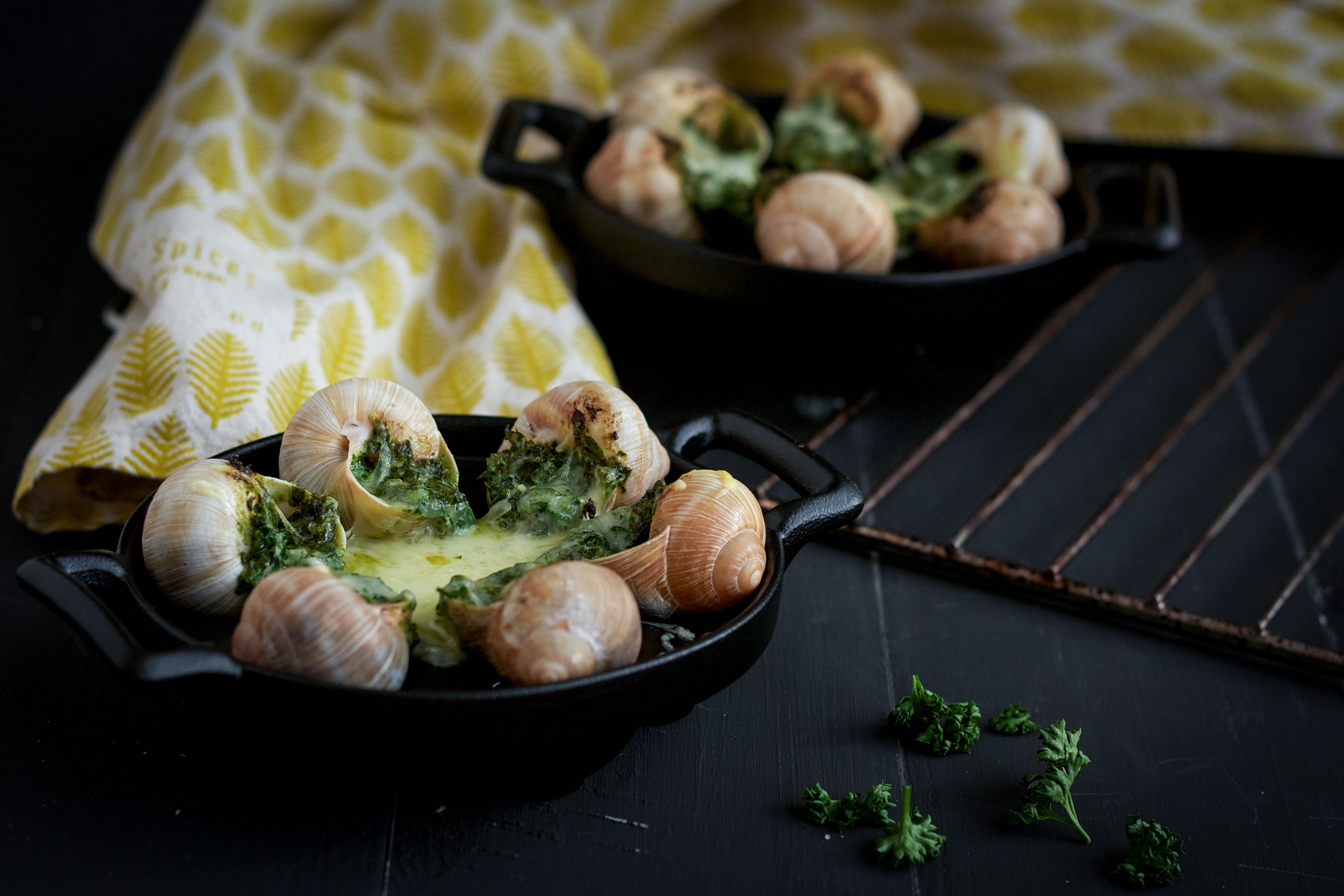 A black cast iron skillet with snails and broccoli on a dark surface.