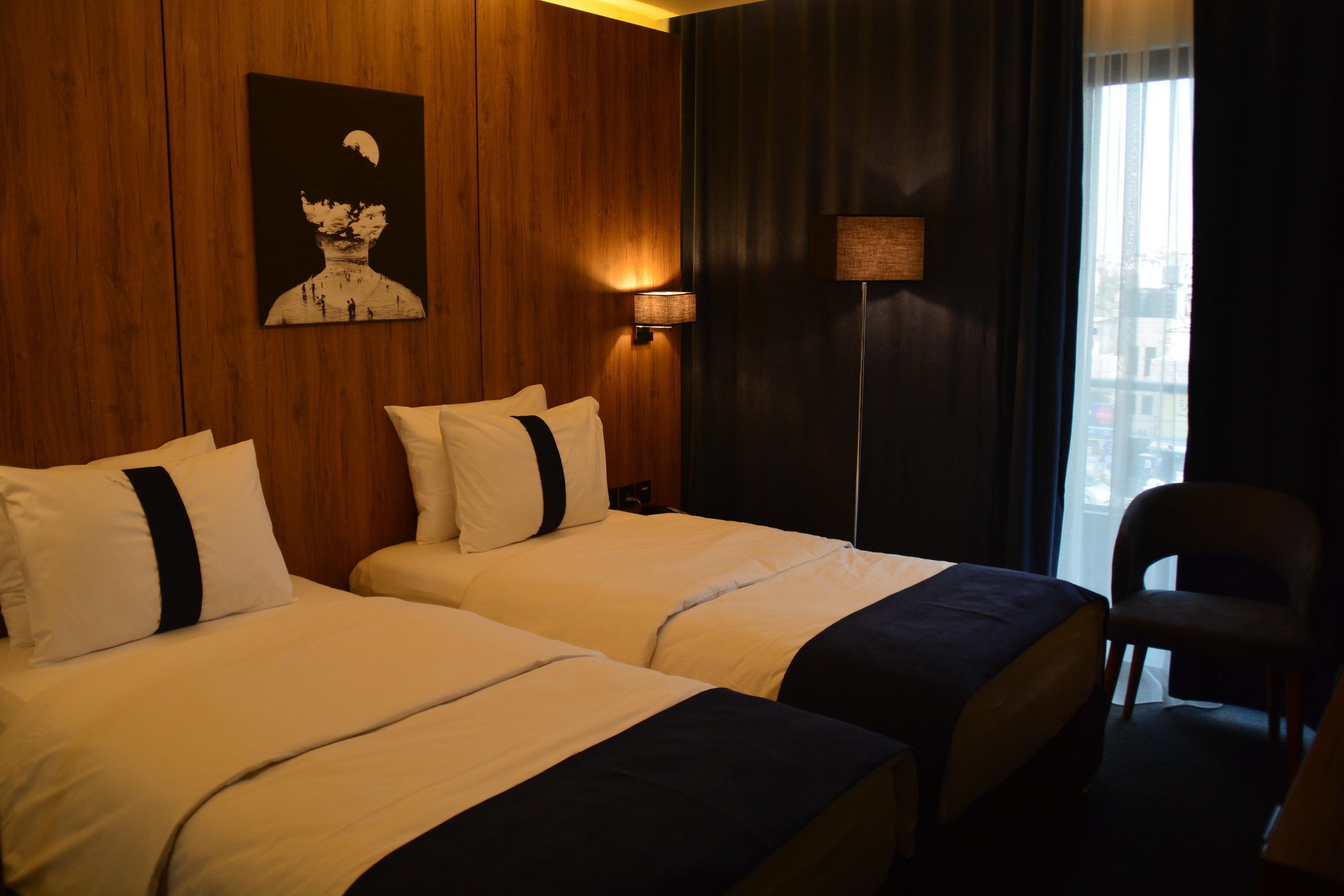 A hotel room with two beds, a wall-mounted artwork, a floor lamp, and a view of the city through a window.