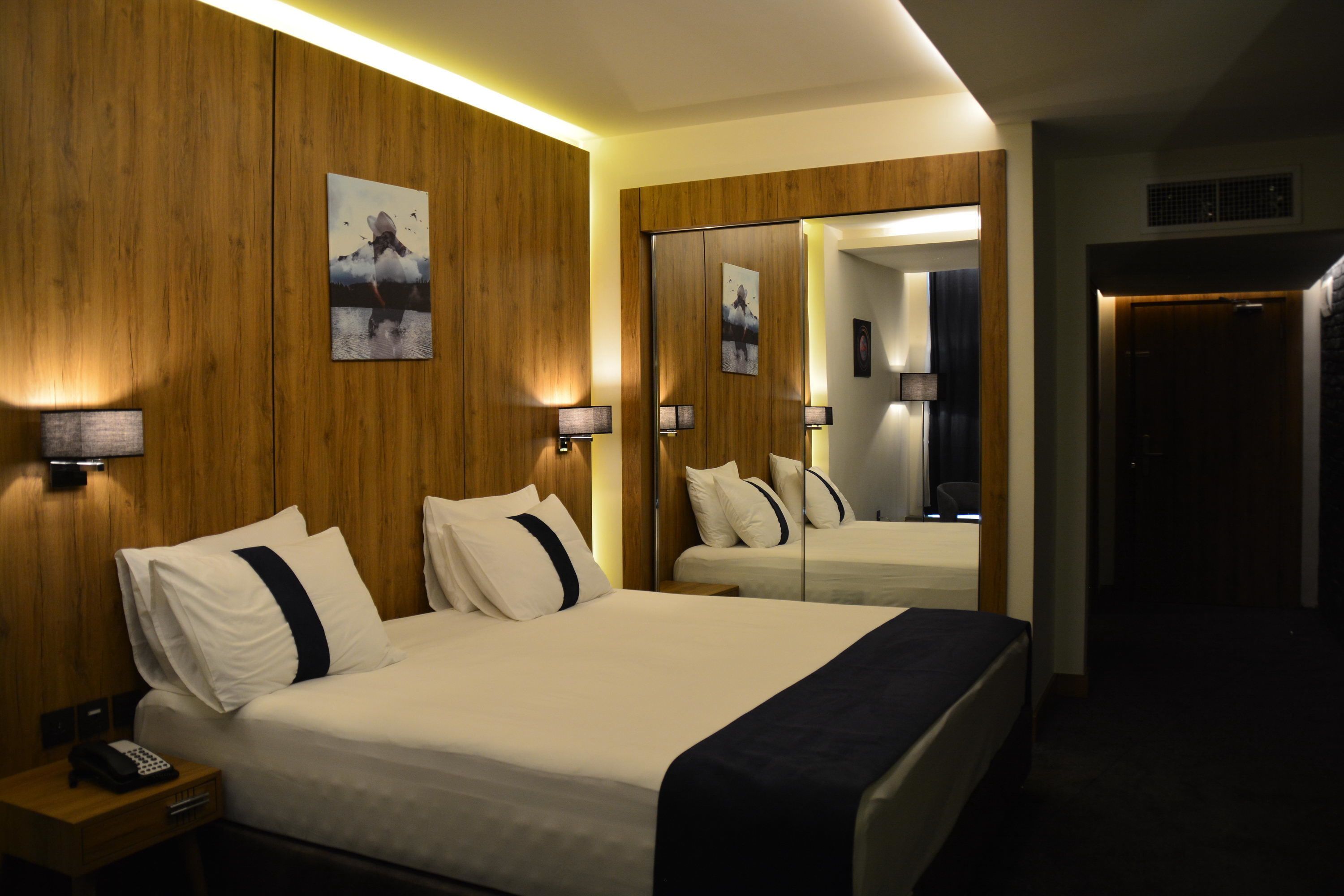 A hotel room with a large bed, bedside lamps, a wooden wall, and a mirror reflecting the room.