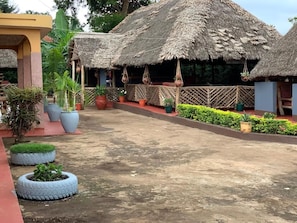 A courtyard with thatched-roof huts, potted plants, and a paved walkway. - The Better Inn (Moshi)